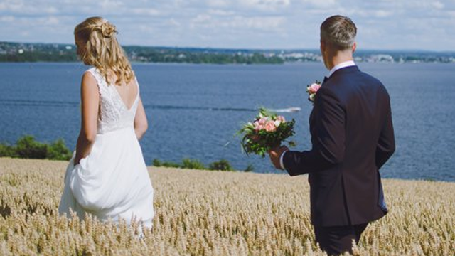 Brudeparet går i en kornmark med utsikt over vannet, bruden i hvit kjole og brudgommen med blomster.