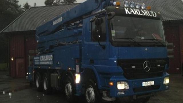 Blå lastebil fra Karlsrud med kran, parkert foran en rød bygning på en grå dag.
