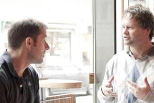 To menn som sitter i en kafé og har en samtale, med fokus på ansiktsuttrykk og kroppsspråk.