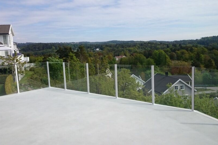 Utsikt fra en moderne terrasse med glassrekkverk, omringet av grønne trær og landskap.