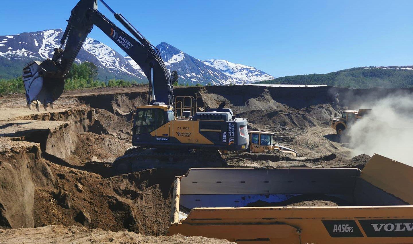 Gravemaskin som arbeider på en byggeplass med snødekte fjell i bakgrunnen. Støv og jord er synlig.