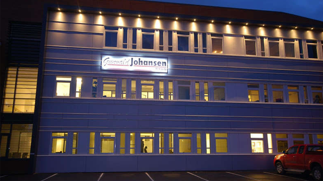 Bygning med opplyst fasade og skilt med navnet "Johansen" i kveldsmørket. Parkeringsplass foran bygningen.