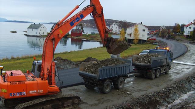 Gravemaskin laster stein i en lastebil ved kysten, med hus og sjø i bakgrunnen.