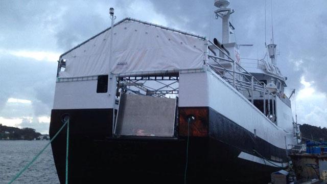 Fiskebåt med hvit presenning og mørk bunn, fortøyd ved havnen under en overskyet himmel.