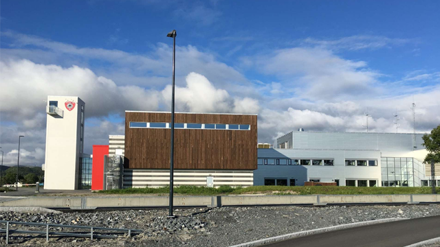 Moderne bygning med tre forskjellige fasader, plassert under en blå himmel med skyer.