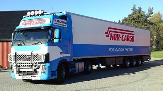 En stor lastebil med Nor-Cargo-logoen, parkert på en gårdsplass med bygninger i bakgrunnen.