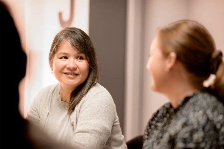 To kvinner i samtale under et møte, med fokus på den smilende kvinnen i grå genser.