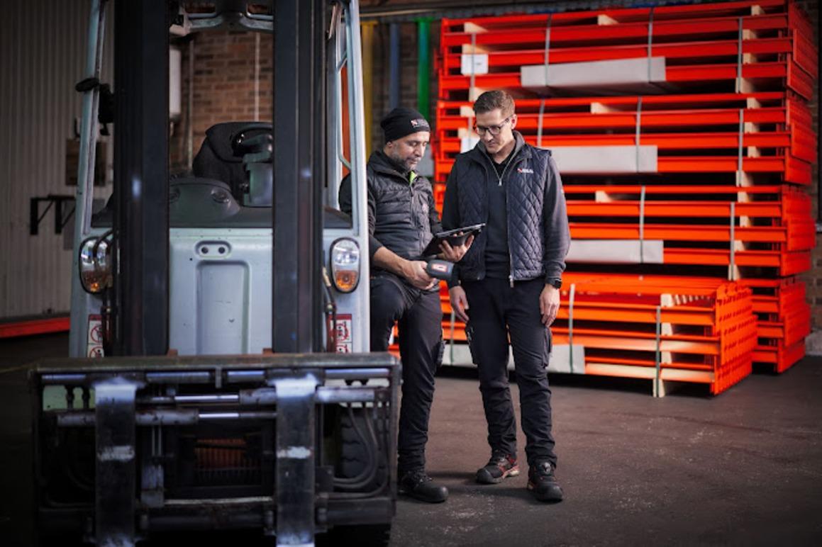 To personer som ser på en nettbrett ved en gaffeltruck i et lager med stabel av paller i bakgrunnen.