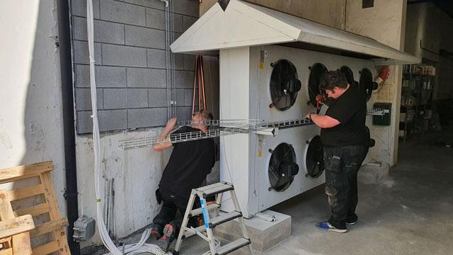 To personer jobber med installasjon av kjølesystem på en vegg, med store vifter synlige.