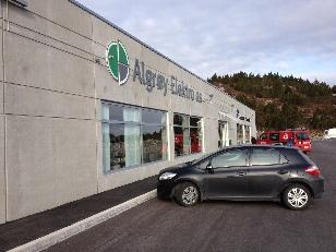En grå bygning med skiltet "Alegro Elektro AS" og en svart bil parkert foran.