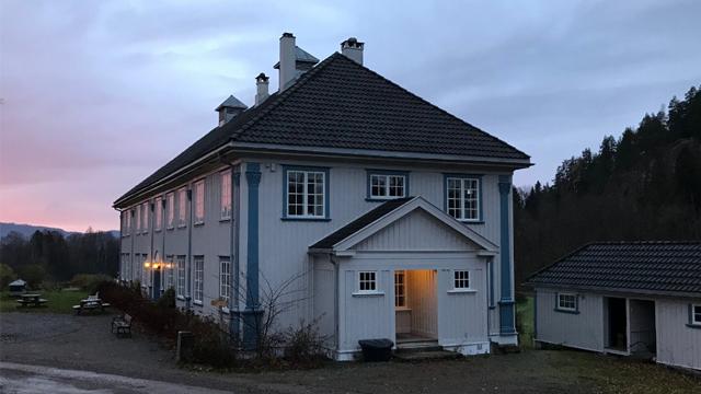 Et stort, hvitt hus med blå detaljer og takvinduer, omgitt av natur i skumringen.