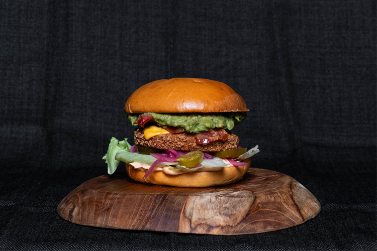 En saftig hamburger med guacamole, salat, ost og syltet rødløk på en treplate.