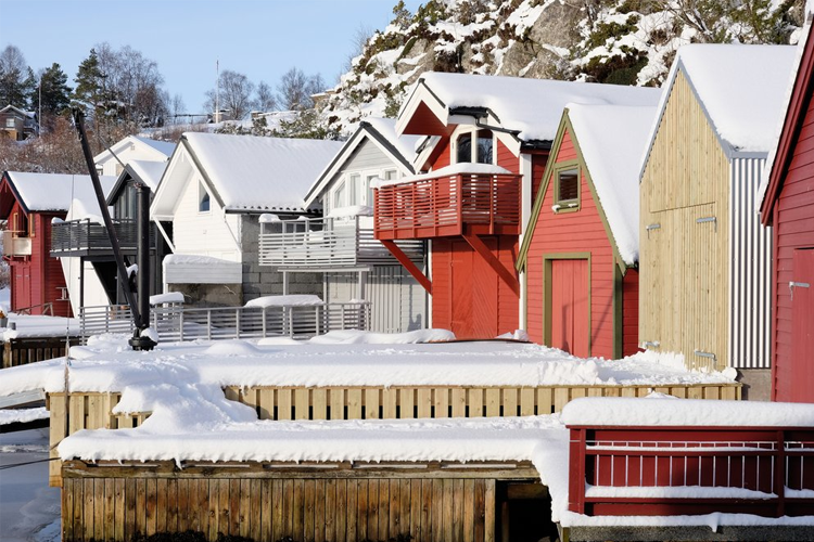 Fargerike hytter med snø på takene, ved en brygge i en vinterlig kystlandsby.
