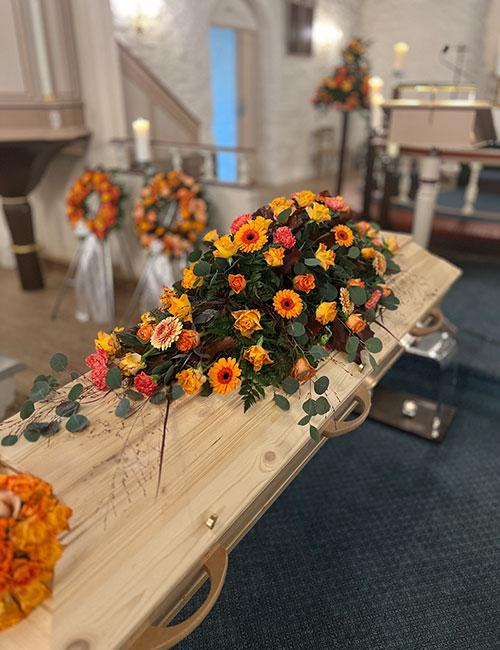En kiste dekorert med oransje og gule blomster i en kirke, med lys og blomsteroppsatser i bakgrunnen.