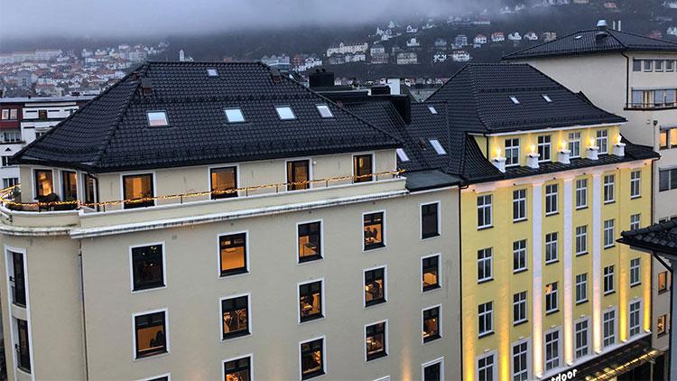 Utsikt over bygninger med takvinduer og belysning i Bergen, med fjell i bakgrunnen.