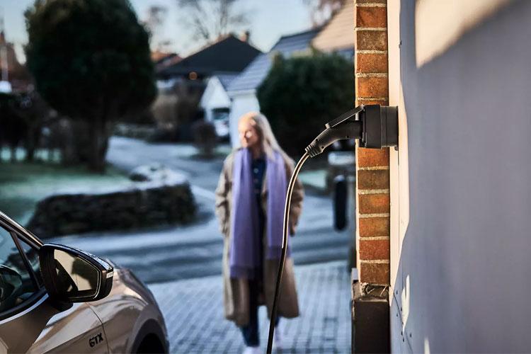 En kvinne i en lang frakk går forbi en elbil som lades ved en murvegg i et boligområde.