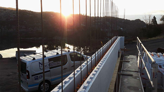 Byggeplass med en hvit varebil, stålstenger og soloppgang over vannet.