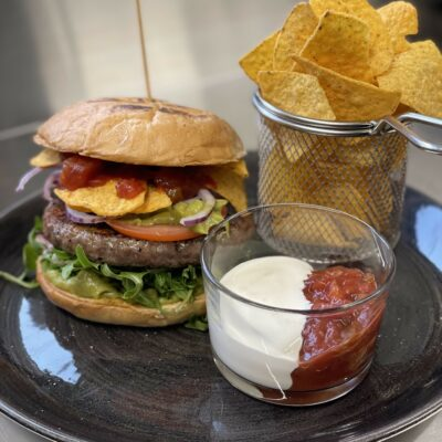 Saftig hamburger med salat, løk, nachos og salsa, servert med chips og rømme i glass.