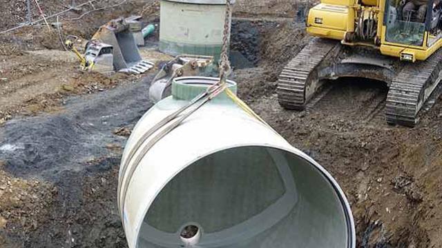Kran som løfter en stor betongrørdel på en byggeplass med gravemaskin i bakgrunnen.
