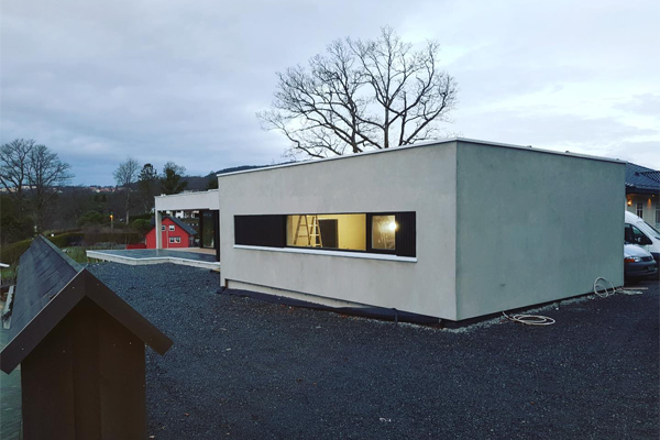 Moderne enebolig med stor vindu, plassert på gruslagt område med trær i bakgrunnen.