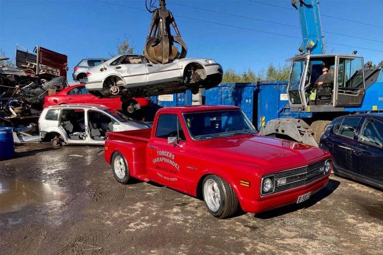 Rød pickup truck i en bilskrapgård med en heis som løfter en bil over den. Skrapbiler i bakgrunnen.