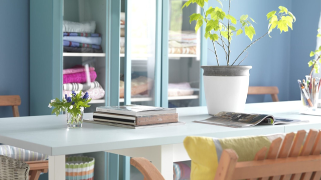 Lyst og moderne rom med et hvitt bord, stoler, planter og hyller fylt med tekstiler.