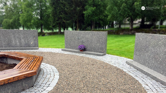 Minnested med grå steinvegger, en blomsterkrans og en trebenk i en grønn park.