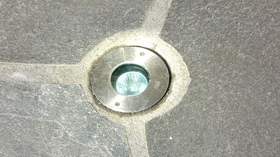 Lysinnretning i bakken med metallramme og glasslinse, plassert mellom steinplater.