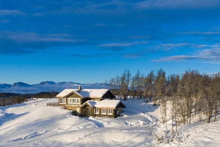 Et koselig hytte i snødekt landskap med fjell i bakgrunnen og klar blå himmel.