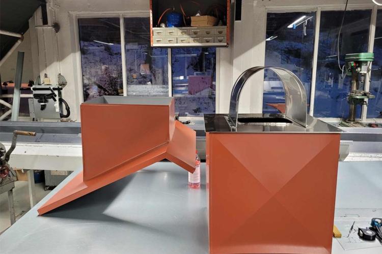 To store og firkantede metallbokser i oransje og sølv, plassert på et arbeidsbord i en verksted.