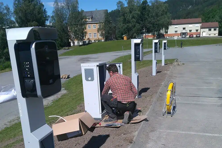 En arbeider installerer ladestasjoner for elbiler på en parkeringsplass med bygninger i bakgrunnen.