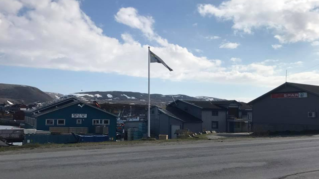 Bygninger med flagg i en fjellandsby under en skyet himmel. Snø på fjellene i bakgrunnen.