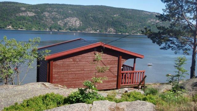 Hytte ved vannet med rød fasade, omgitt av steiner og trær, med fjell i bakgrunnen.