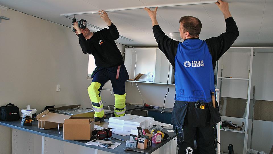 To personer som installerer takplater i et rom, med verktøy og materialer spredt rundt.