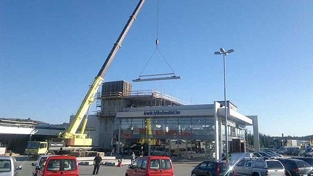 Kran løfter en stor metallramme over en bygning under konstruksjon på en parkeringsplass.