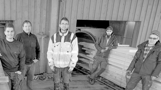 Fem menn i arbeidsklær står i en trebearbeidingsfabrikk med treplanker i bakgrunnen.