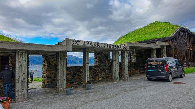 Inngangen til Brekkestranda med grønt tak og steinvegg, en bil parkert foran bygningen ved vannet.