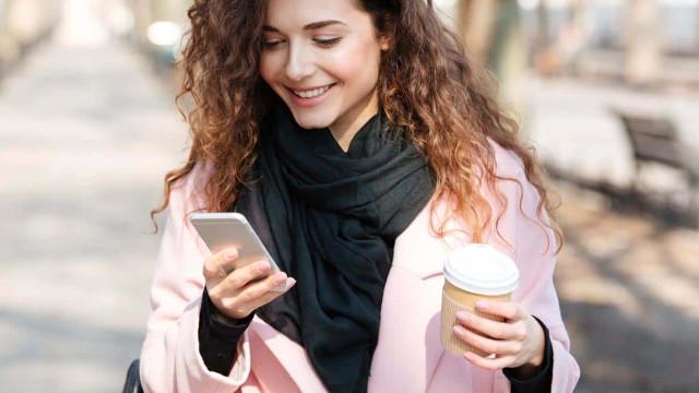 En smilende kvinne med krøllete hår som holder en kaffekopp og ser på telefonen sin i en park.