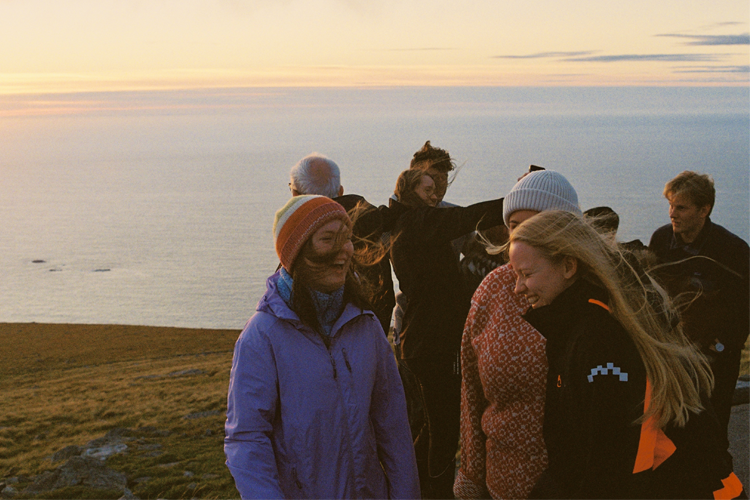 Gruppe mennesker smiler og ler sammen på en fjelltopp med havutsikt under solnedgang.
