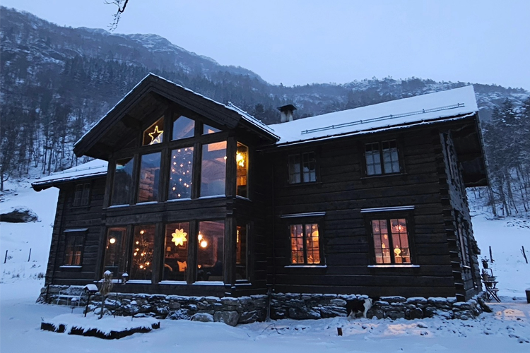 En koselig hytte i snøen med store vinduer og juledekorasjoner, omringet av fjell og trær.