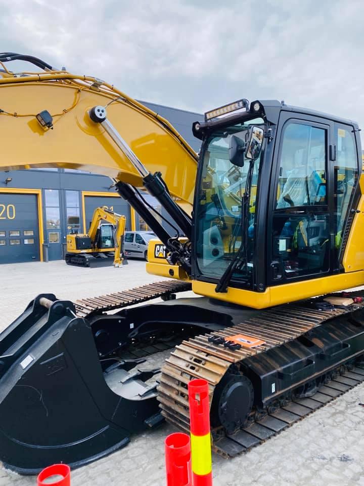 Gravemaskin med gule og svarte detaljer, stående på en byggeplass med andre maskiner i bakgrunnen.
