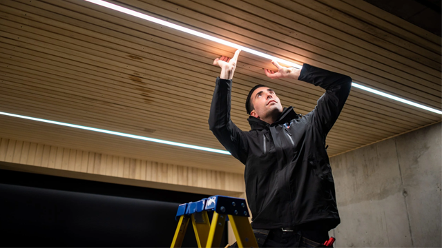 En elektriker står på en stige og installerer takbelysning i et moderne rom med trepanel.