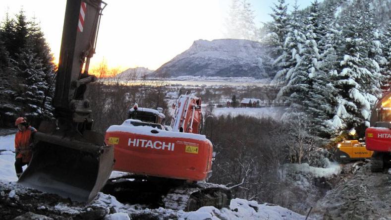 Gravemaskin fra Hitachi i snødekt landskap med fjell og trær i bakgrunnen. Solnedgang gir et varmt lys.