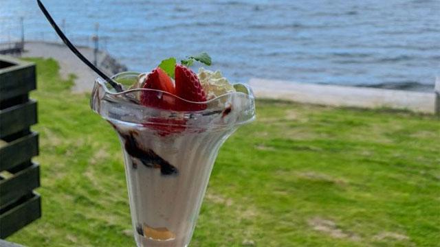 En iskremparfait med jordbær og sjokoladesaus, plassert ved vannkanten med grønt gress i bakgrunnen.