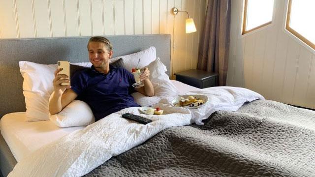 En person sitter i sengen med en telefon i hånden, et glass vin og snacks på en tallerken.