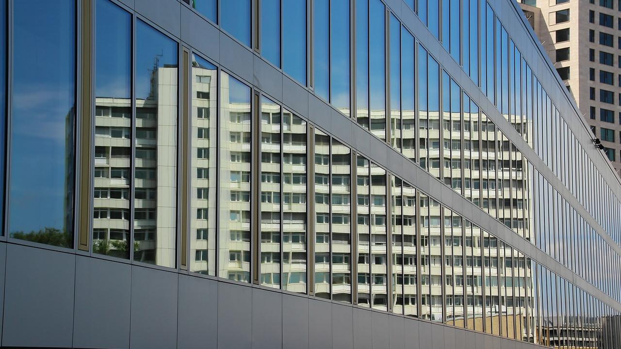 Moderne bygning med speilende glassfasade som reflekterer en boligblokk i bakgrunnen.