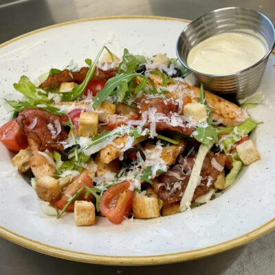 En salat med kylling, bacon, tomater, krutonger og parmesan, servert med en dressing i en liten skål.