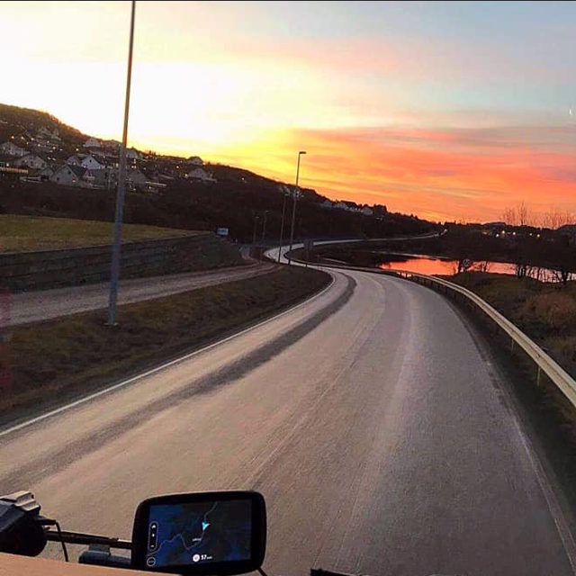 En vei som svinger langs en elv med solnedgang i bakgrunnen og hus på åsene.