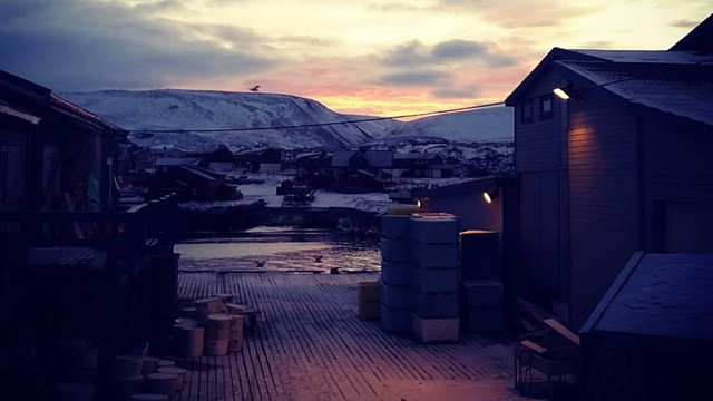 Utsikt over en snødekt landsby ved vannet, med fjell i bakgrunnen og en fargerik solnedgang.