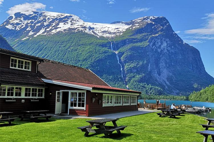 Utsikt over en hytte ved fjorden med fjell og fossefall i bakgrunnen, omkranset av grønt gress og benker.